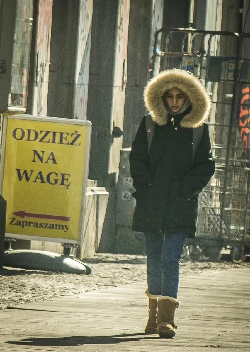 Person in a fur-trimmed hood walking toward a yellow street sign
